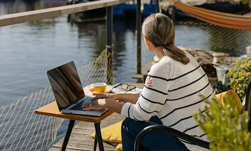 En person sitter med en laptop på en solig brygga vid vattnet.