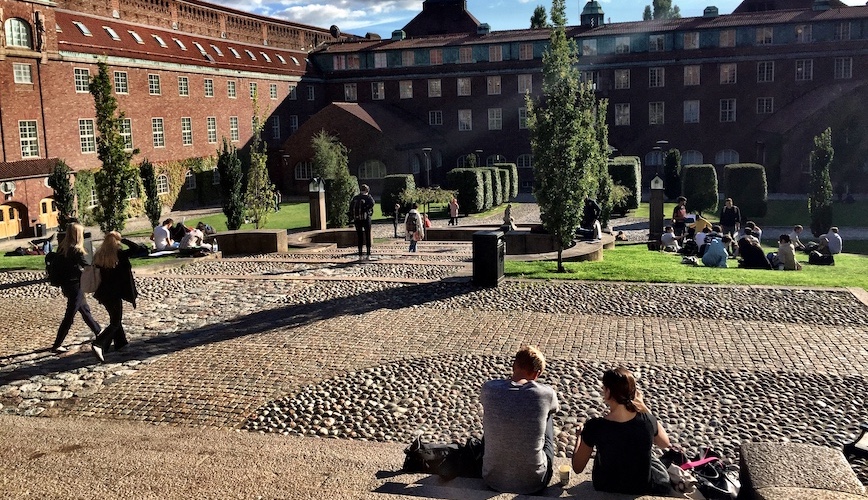 Foto: studenter inne på borggården till KTH:s huvudbyggnad under blå himmel med vita moln.