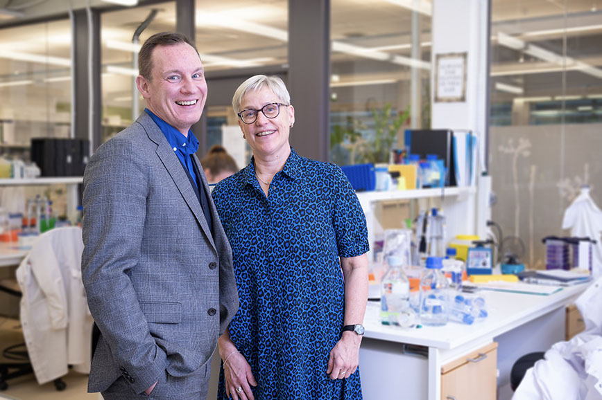 A man and a woman standig in a research lab. Behind them there are bottles on a table.