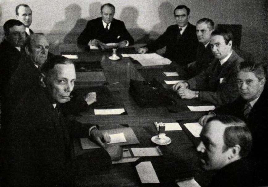 A black and white photo of men with black suits sitting at a table.