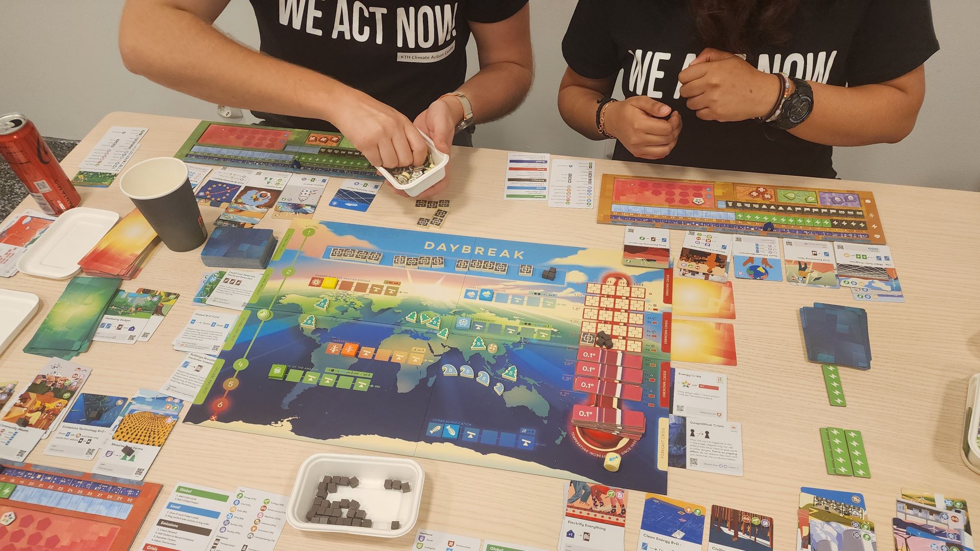 Two students playing the game Daybreak, with a board and cards covering the table. 