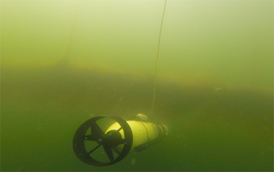 a yellow tube shaped vehicle with a propeller on its tale moving through underwater murk and seaweed